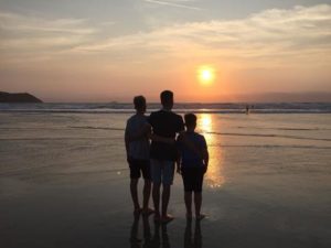 three teenage boys arm in arm looking out at a sunset over a beach
