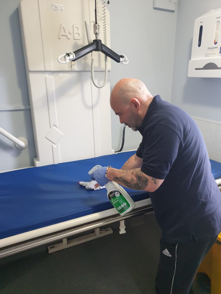 A cleaner using antibacterial spray to clean a medical bed in a social care setting.