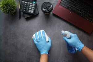 gloved hands viewed from above cleaning a desk with a laptop and phone.