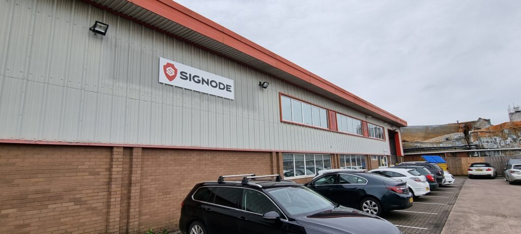 Photograph looking towards the entrance of a grey corrugated industrial building with a sign that reads “Signode”, with a number of cars parked in front, and old brick buildings beyond the car park