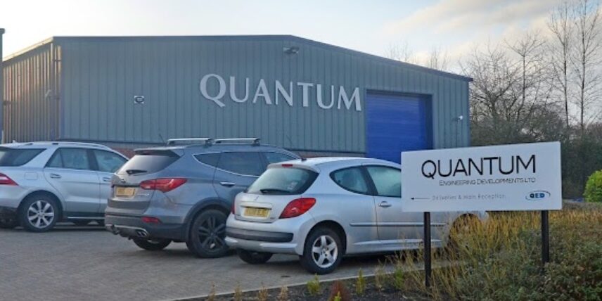 Photograph of the Quantum primacies, showing a grey industrial unit, with a large blue roller shutter in the background with the word "Quantum" on it, with a carpark in the foreground, with three cars parked behind a sign of saying "Quantum"