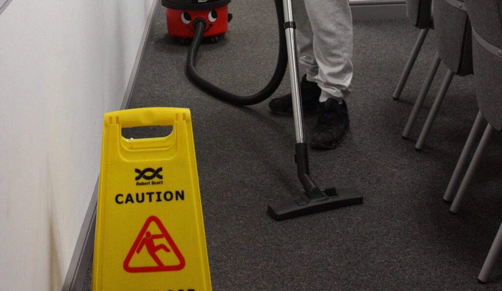 a vacuum cleaner cleaning a carpeted floor next to a yellow caution sign.