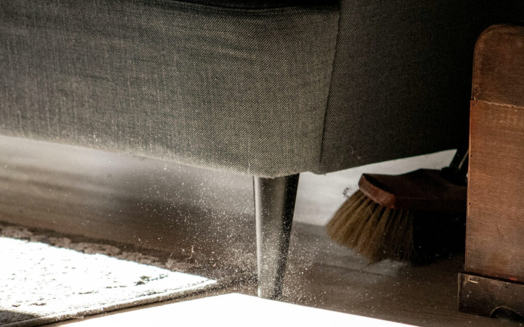 Detail of a brush conducting commercial floor cleaning under a sofa with dust particles blowing up into the air.