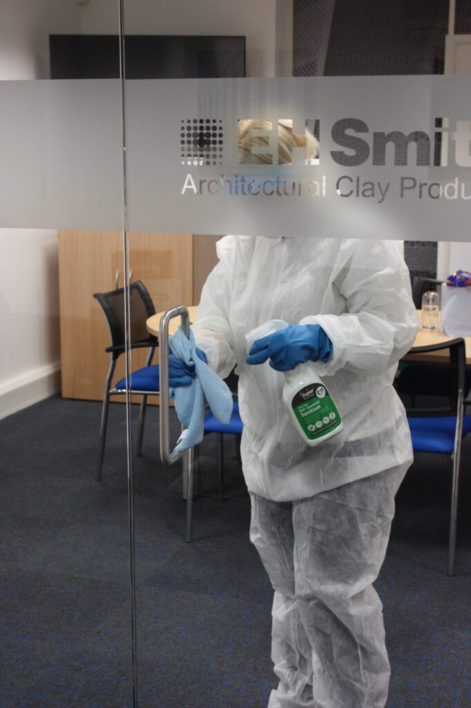 A cleaning opertive stands inside an office with glass walls, wiping down the handle of the door with a blue cloth whilst wearing full body clothing protector