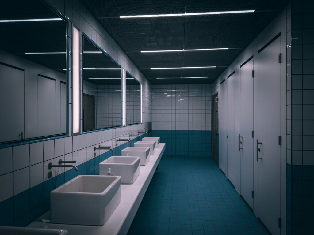 A tiled, white and blue public bathroom with four toilet cubicals and five sinks.