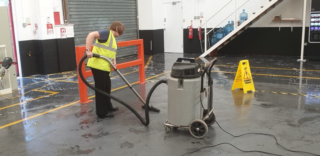 A cleaning operative hoovers water from a warehouse floor with a yellow hazard sign next to them