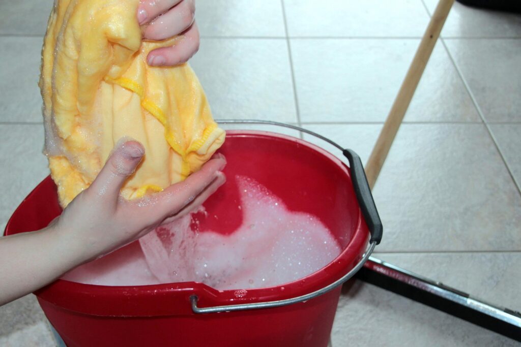 A detail of someone squeezing water out of a yellow cloth over a red bucket full of soapy water